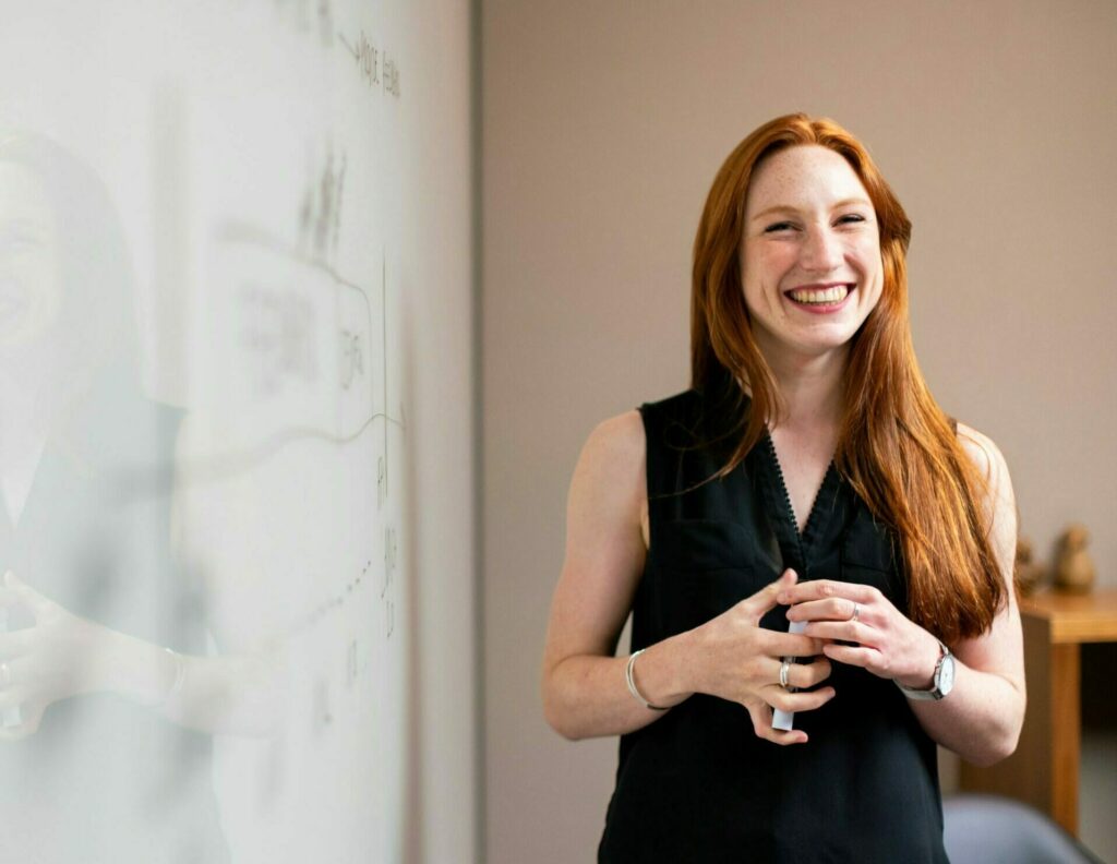 A person smiles while standing near a whiteboard, holding a marker in hand, in a classroom or office setting. The whiteboard features faint, indistinct sketches or diagrams.